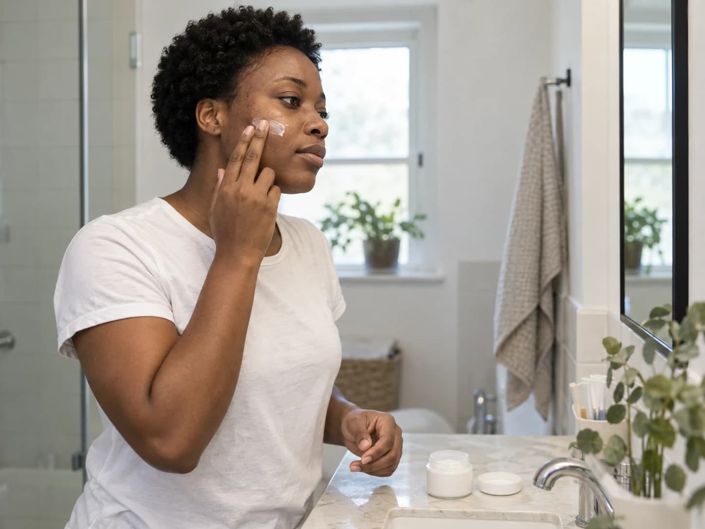 A quiet moment of skincare: a woman applying moisturizer at her bathroom mirror.